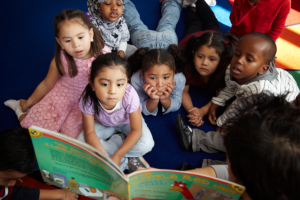 Children sit together and listen as a teacher reads a book aloud during story time.