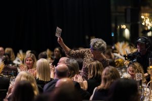 Woman holding up an auction paddle at a crowded dinner table.