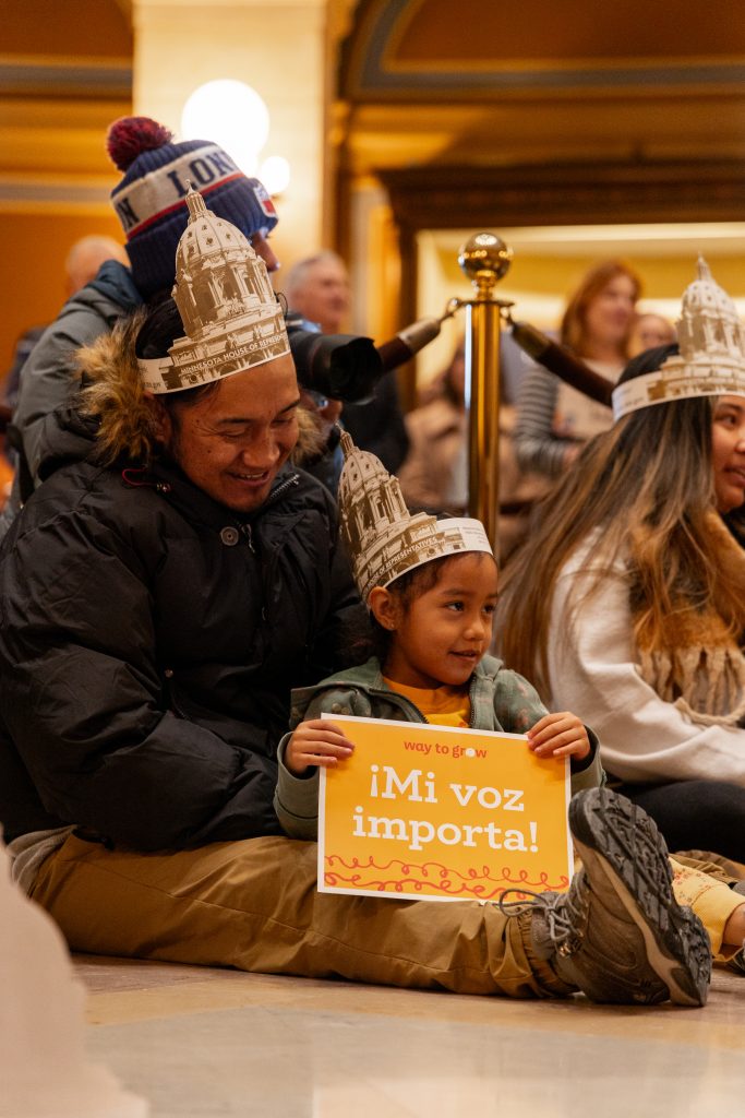 Latino father with his preschool daughter sitting on the floor in the Minnesota State Capitol. They are wearing paper hats depicting the Capitol building and holding a sign that says, "¡Mi voz importa!" and smiling.