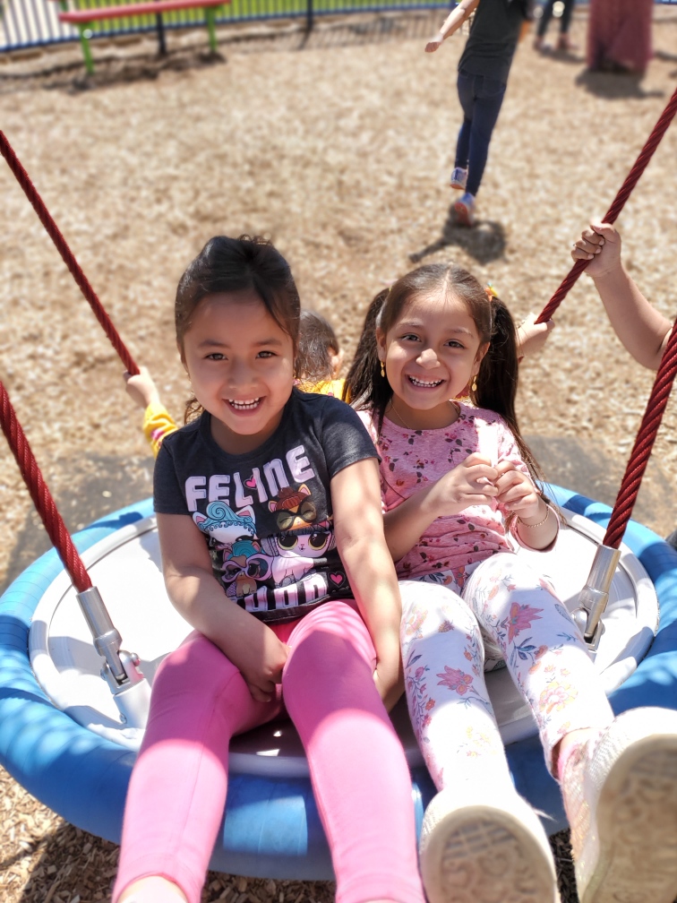 Two preschool girls swinging on a swing at the park.