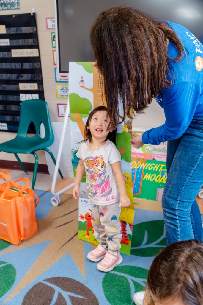 PNC volunteer holds up growth chart and a preschooler measures herself against it.