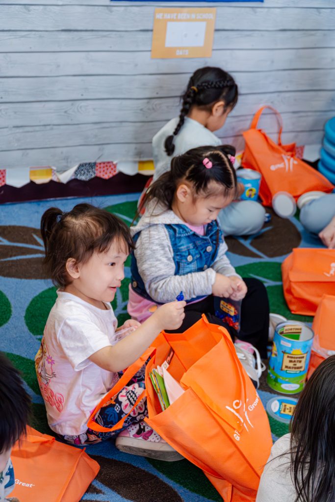 Preschoolers sitting on the floor of their classroom , opening orange bags from PNC, filled with books, math manipulative, and more.