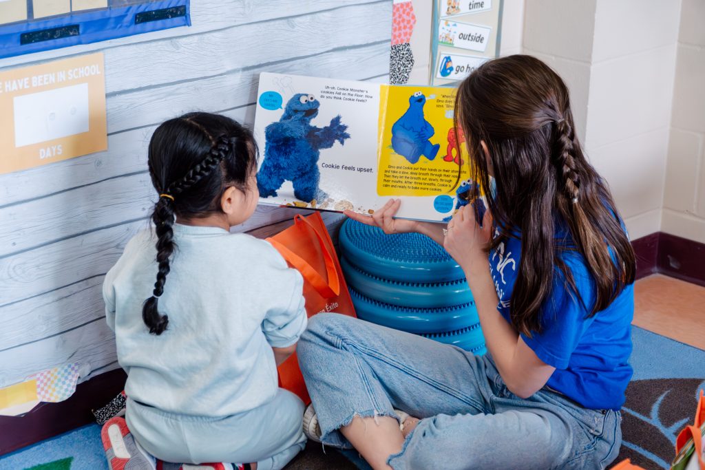 PNC volunteer reads a Sesame Street book to a preschooler on the floor.