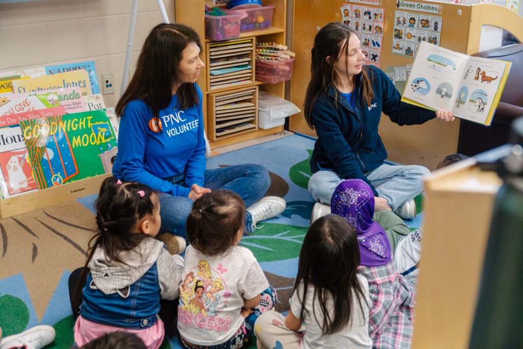 Two PNC volunteers read a story to a group of preschoolers sitting together on the floor.