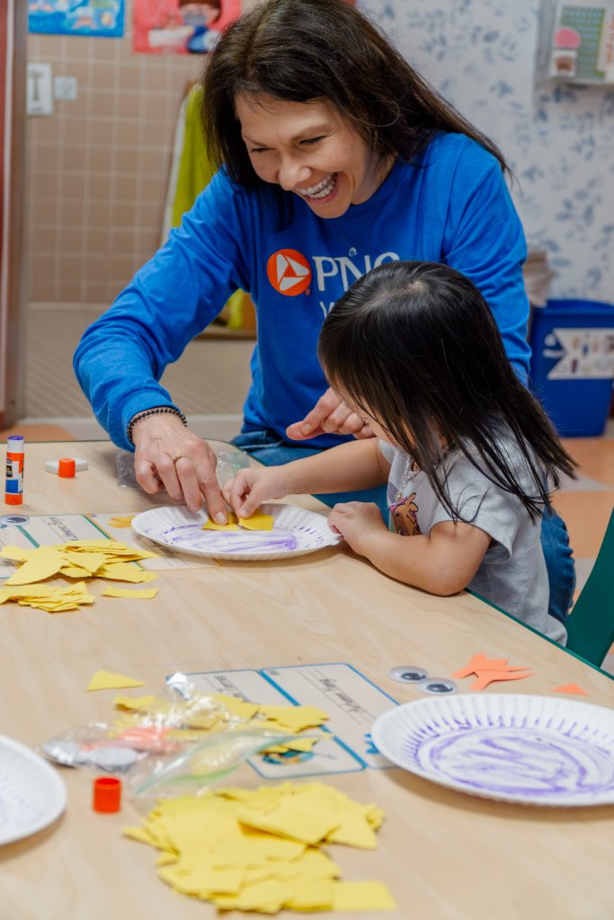 PNC volunteer assists a preschooler in a craft gluing yellow paper to a paper plate, making a baby chick.
