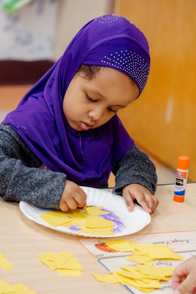 Preschooler in a purple hijab gluing yellow paper on a paper plate to make a chick.