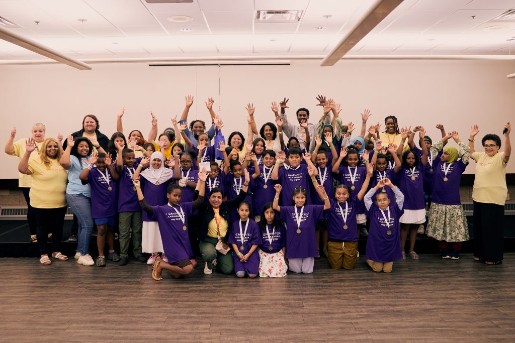 Group photo of third grade graduates and staff, everyone smiling with their hands in the air. Third graders are wearing purple shirts and medals. Staff is wearing yellow shirts with Way to Grow logo.