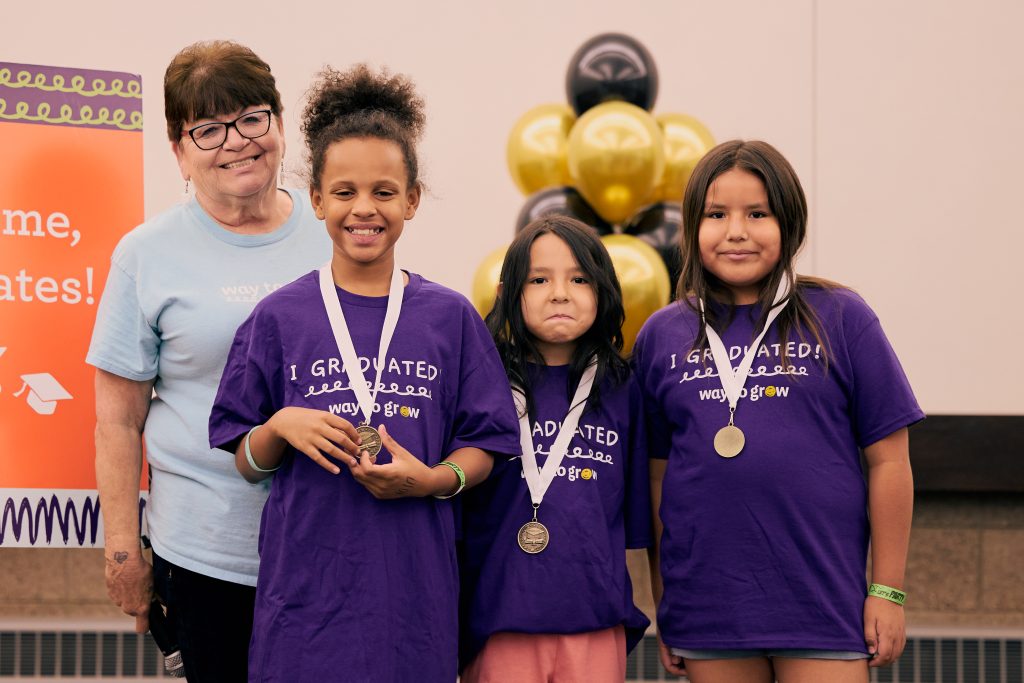 Three third grade graduates in purple shirts and medals, smiling with a Family Educator.