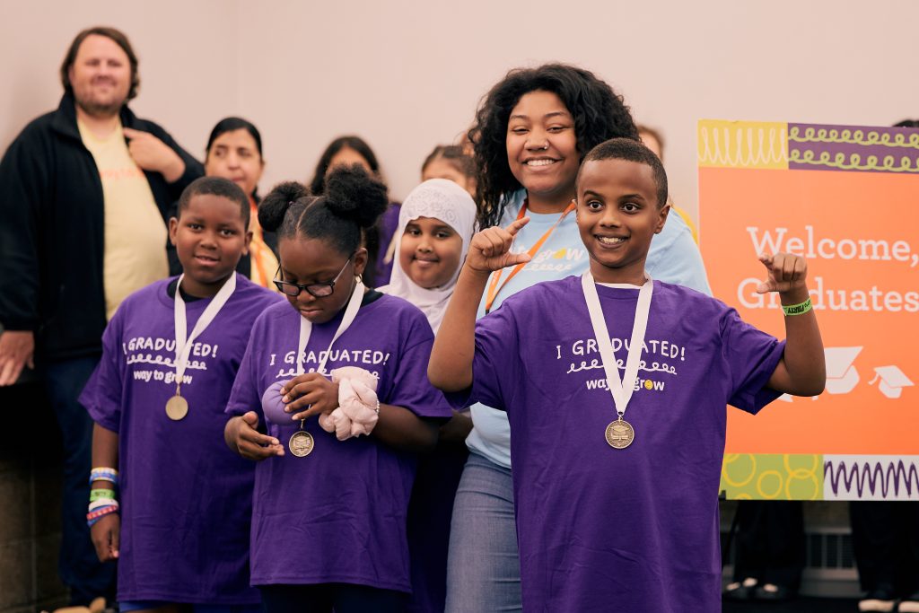 Three third grade graduates in purple shirts and medals with a Family Educator behind them.